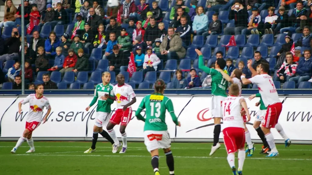 "Jogadores de futebol disputam a bola em campo durante uma partida, com torcida atenta nas arquibancadas ao fundo."