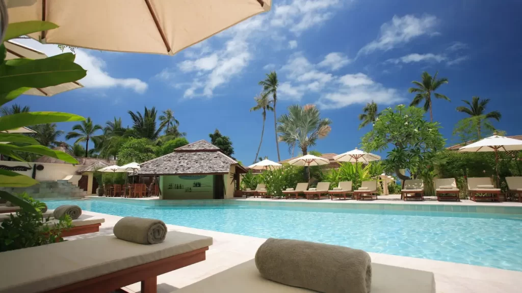 “Piscina de resort cercada por espreguiçadeiras, guarda-sóis e palmeiras sob um céu azul — cenário ideal para férias relaxantes.”