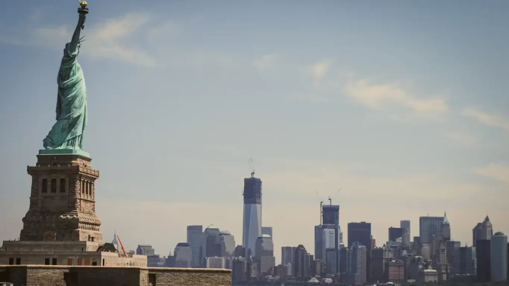 "Estátua da Liberdade em primeiro plano com os arranha-céus de Manhattan ao fundo sob um céu azul claro."