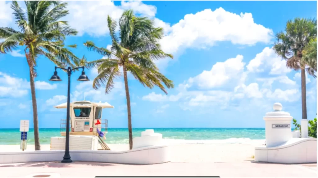 Cabana de salva-vidas na praia de Fort Lauderdale, com palmeiras ao vento e mar azul sob céu com nuvens claras.