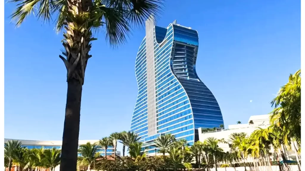 Prédio em formato de guitarra cercado por palmeiras sob um céu azul claro, localizado próximo a praias tropicais.