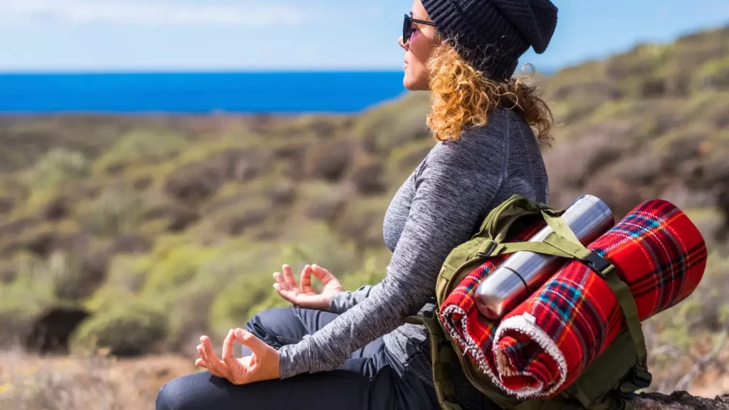 Mochileira sentada na natureza, meditando com mochila e utensílios de acampamento ao fundo.
