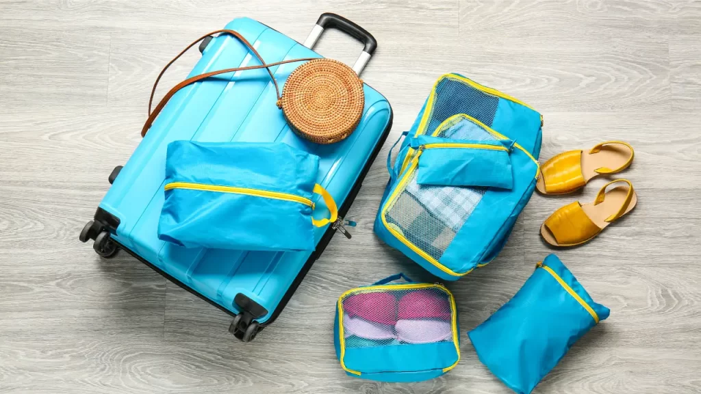 Mala azul com cubos organizadores, bolsa de palha e sandálias prontas para uma viagem de verão.