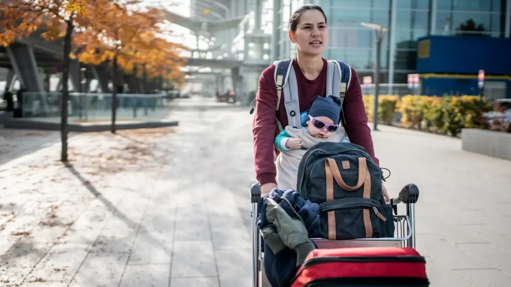 Mãe usando um canguru com um bebê, empurrando um carrinho de bagagem, chegando de viagem em um aeroporto, com árvores de outono e um prédio moderno ao fundo.