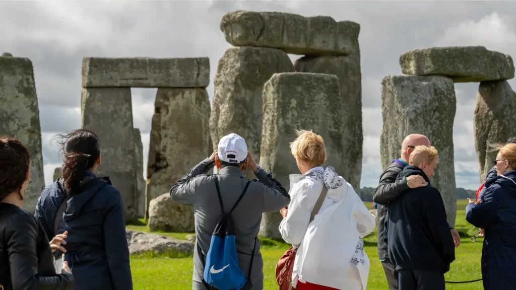 Um grupo de pessoas observando as grandes pedras de Stonehenge, com céu nublado ao fundo e gramado verde ao redor.