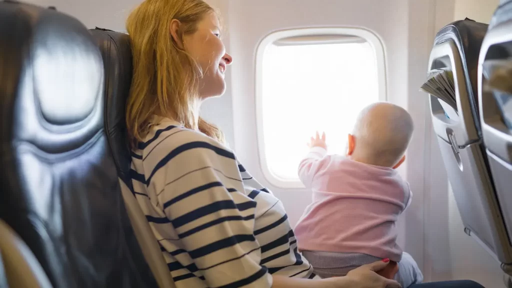 Mãe segurando um bebê no colo dentro de um avião, com o bebê olhando pela janela, ambos em assentos próximos a uma janela iluminada.