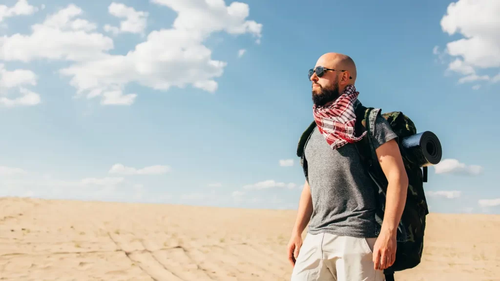 Mochileiro caminhando por uma paisagem desértica sob céu azul, com mochila e lenço no pescoço.