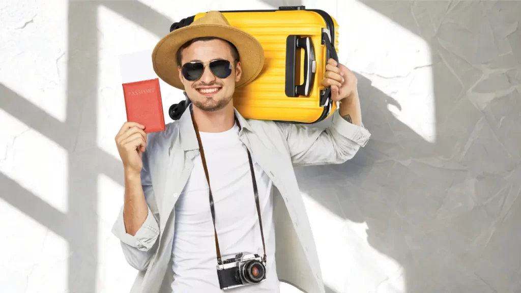 Homem sorridente com chapéu, óculos escuros e câmera no pescoço segura passaporte e mala amarela, pronto para viajar.