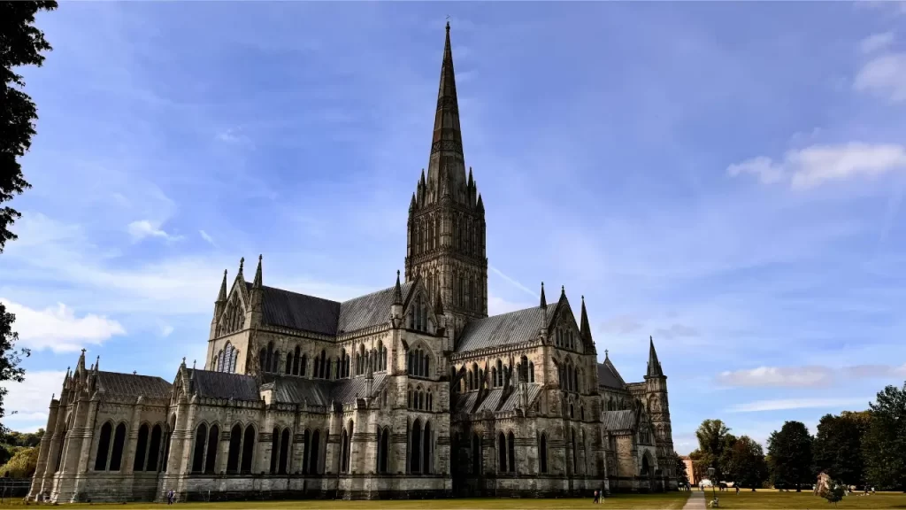 Uma catedral gótica com uma alta torre central e arcos detalhados, cercada por gramado verde e árvores sob um céu azul claro.