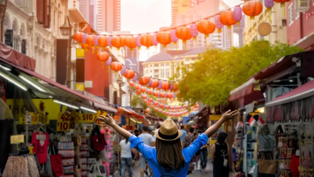 Mochileira com os braços abertos caminhando por mercado de rua decorado com lanternas asiáticas.