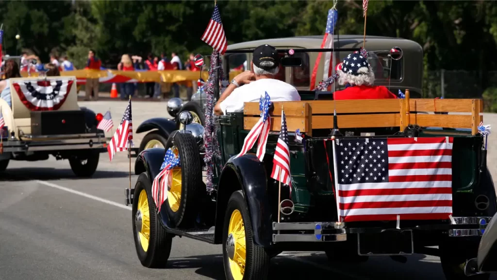 Um carro vintage decorado com bandeiras americanas participa de um desfile do 4 de Julho. Dois ocupantes estão sentados na parte de trás, com bandeiras pequenas e uma bandeira grande pendurada na traseira. Outros carros e pessoas ao fundo também exibem decoração patriótica.