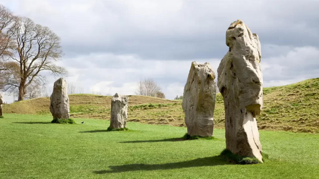 Cinco grandes pedras verticais dispostas em uma colina verdejante, com uma árvore e céu nublado ao fundo.