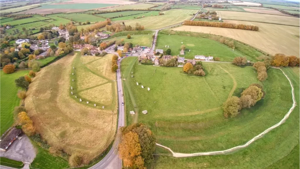 Uma vista aérea de um círculo de terra com pedras dispostas em um campo verde, perto de uma vila e campos agrícolas, com árvores de outono.
