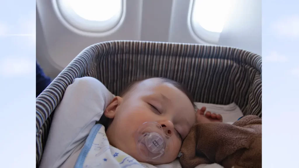 Bebê dormindo em um berço a bordo de um avião, com uma chupeta na boca e um bichinho de pelúcia marrom ao lado, perto de janelas com vista para o céu.