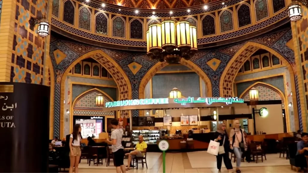 Uma imagem do interior de uma loja da Starbucks Coffee em um shopping com arquitetura árabe detalhada. O teto é ornamentado com arcos, azulejos coloridos em tons de azul, amarelo e branco, e lustres de estilo tradicional. O balcão da Starbucks exibe o logotipo verde, com clientes sentados e caminhando ao redor, segurando sacolas de compras. Há uma placa com texto em árabe e inglês, e o ambiente é bem iluminado.