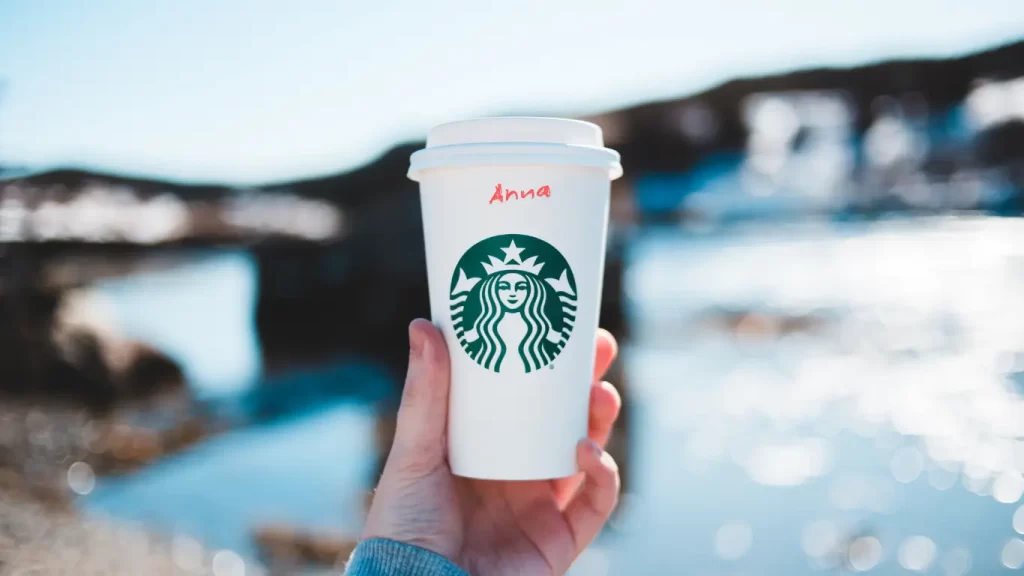 Copo do Starbucks com o nome “Anna” escrito à mão, segurado por uma pessoa ao ar livre com paisagem desfocada ao fundo.