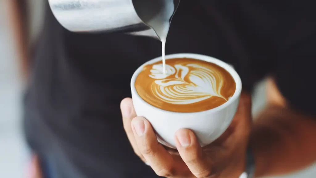 Uma imagem de close-up mostrando uma mão segurando uma xícara branca de café latte, com leite sendo despejado de um jarro de metal por outra mão. O leite forma um desenho em forma de coração e ondas na superfície cremosa do café, destacando a habilidade de um barista. O fundo está desfocado, focando na xícara e no processo de preparo.