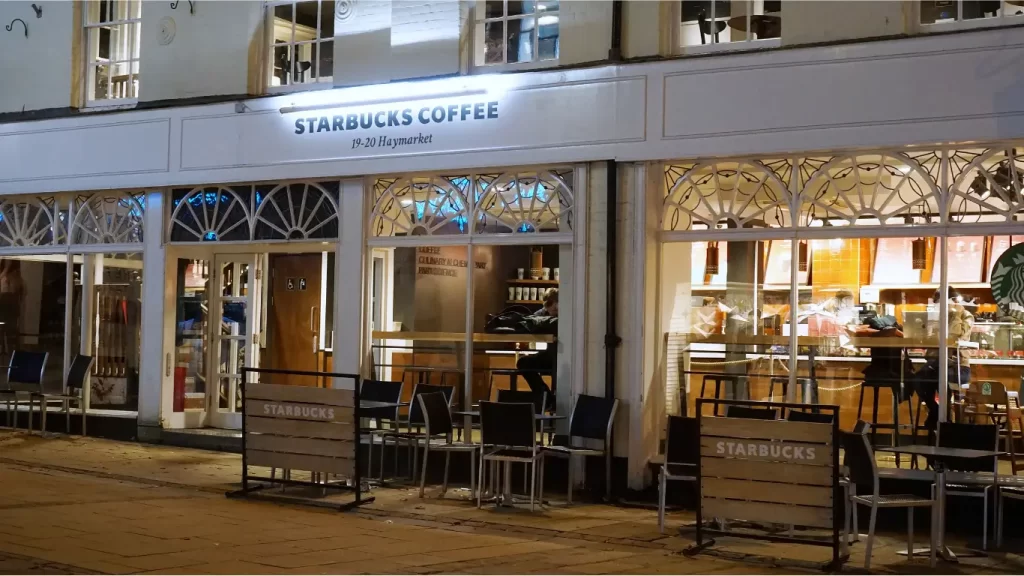 Uma foto noturna de uma loja da Starbucks Coffee localizada no número 19-20 Haymarket. A fachada da loja exibe grandes janelas arqueadas com detalhes ornamentais, iluminadas por dentro, revelando o interior acolhedor com mesas, cadeiras e o logotipo da Starbucks. Há algumas mesas e cadeiras vazias na calçada em frente à loja, protegidas por barreiras com o nome "Starbucks". A iluminação destaca a arquitetura clássica do prédio e cria uma atmosfera convidativa.